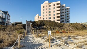 A pathway leading to a modern building with multiple floors, surrounded by sand dunes and vegetation. - Sound of the Sea 203N - 2BR Condo w/ Pool! (Emerald Isle)