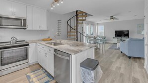 A modern kitchen with white cabinets, a built-in oven, and a dishwasher. There is a wooden spiral staircase leading to a dining area with a TV and a blue sofa. - Sound of the Sea 203N - 2BR Condo w/ Pool! (Emerald Isle)