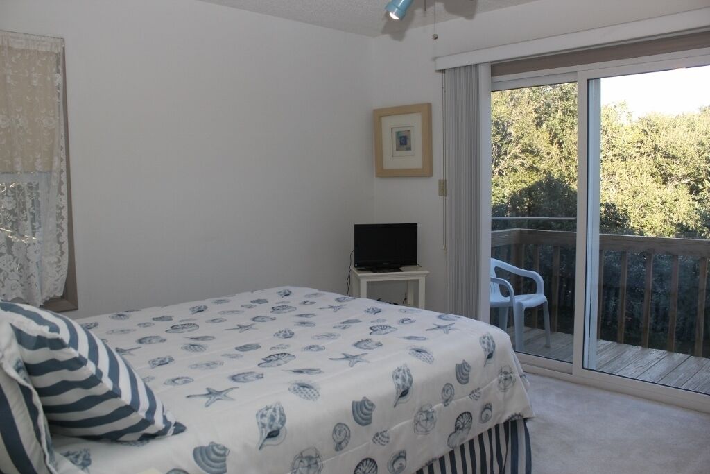 Une chambre avec un lit, un téléviseur posé sur une petite table, un tableau encadré au mur et une porte-fenêtre coulissante donnant sur un espace extérieur avec une terrasse en bois et une chaise.