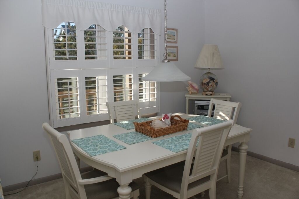Un coin repas avec une table et des chaises blanches, une lampe suspendue et une fenêtre avec des volets.