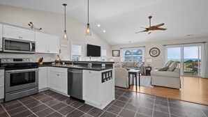 A modern kitchen with white cabinets, a built-in oven, and a dishwasher. There is a dining area with a table and chairs, and a large window offering a view of the ocean. - POOL & DOGS WELCOME! Easy Beach Access (Emerald Isle)