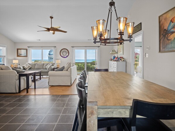 A spacious living area with a wooden dining table, two sofas, a ceiling fan, and a view of the ocean.