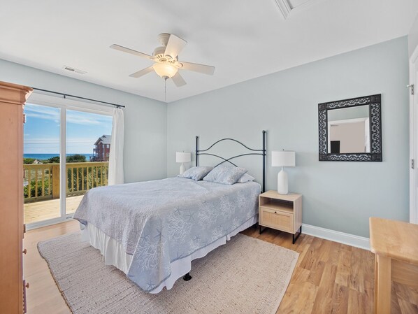 Ein Schlafzimmer mit Bett, Nachttischen, Deckenventilator, einem großen Fenster mit Blick auf den Strand und einem Spiegel an der Wand.