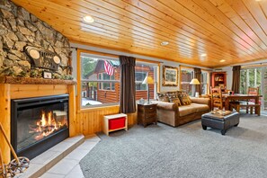 A cozy living room with a fireplace, wooden walls, and large windows.