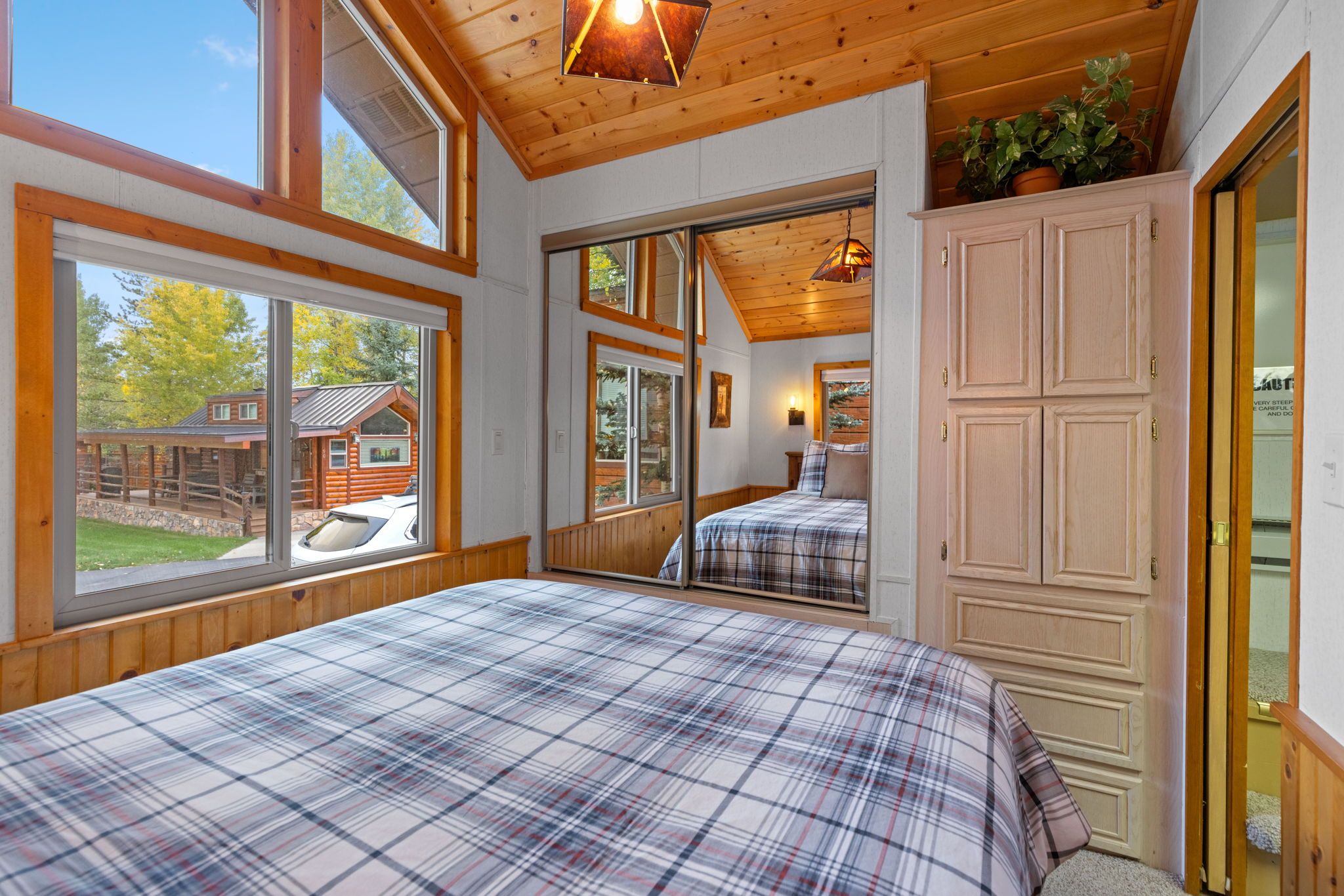 A bedroom with a large bed, wooden ceiling, and a view of a cabin.