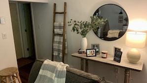 En hyggelig stue med sofa, en træstige, et spejl, en vase med grønne planter, et lille bord med bøger og stearinlys, samt en lampe.