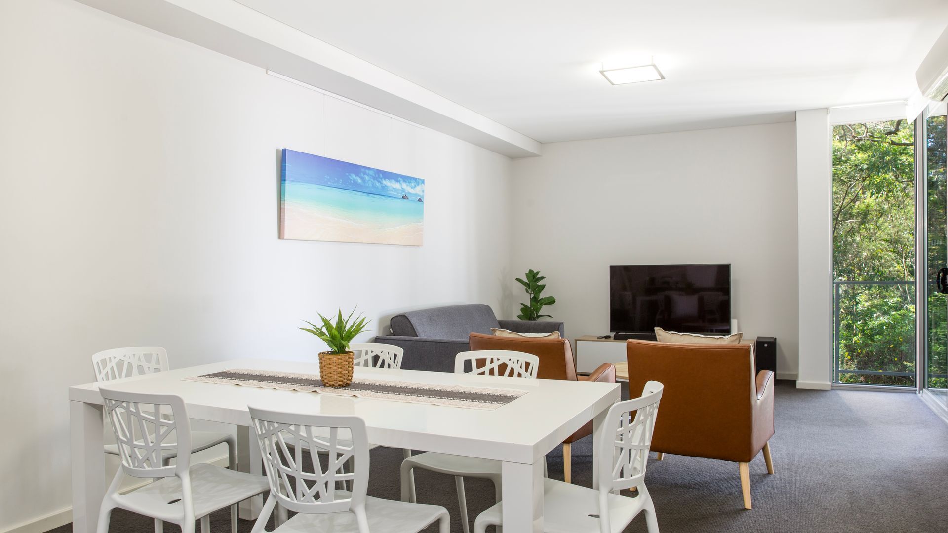 A modern living room with a white dining table, white chairs, a grey sofa, a brown armchair, a flat-screen TV, and a large window with a view of trees.