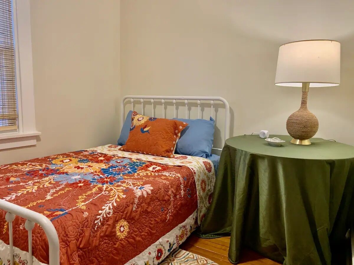 A bedroom with a bed, pillows, a bedside table with a lamp, and a window with blinds.