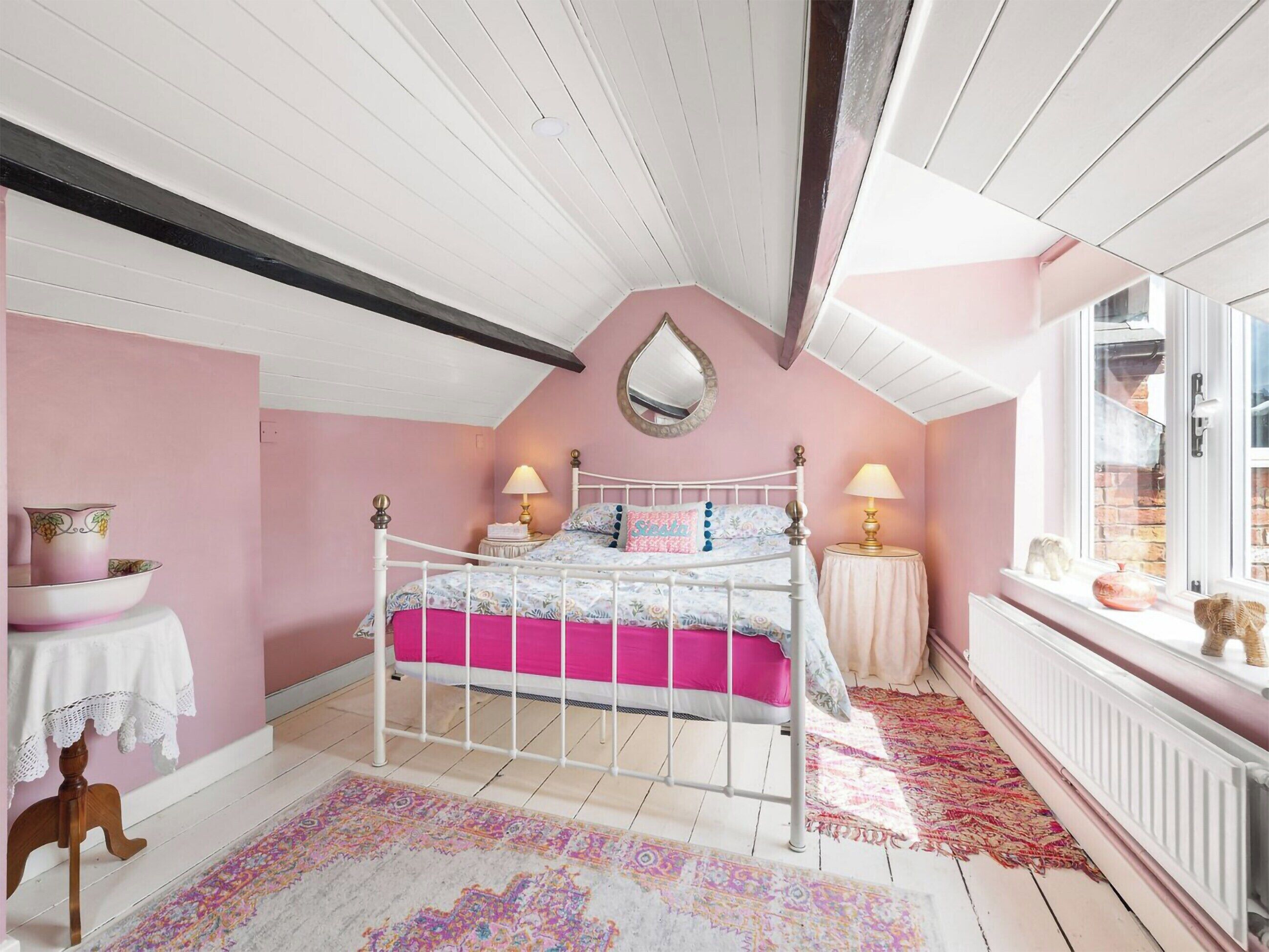 Ein Schlafzimmer mit einem weißen Metallbett, rosafarbenen Wänden, einem großen Spiegel und einem Fenster mit Blick auf ein Backsteingebäude.
