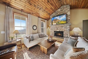 A cozy living room with a stone fireplace, wooden ceiling, and comfortable seating. - Luxury Mountain Retreat with Grandfather Mountain Views | Sleeps 16 | Game Room | Chef's Kitchen (Seven Devils)