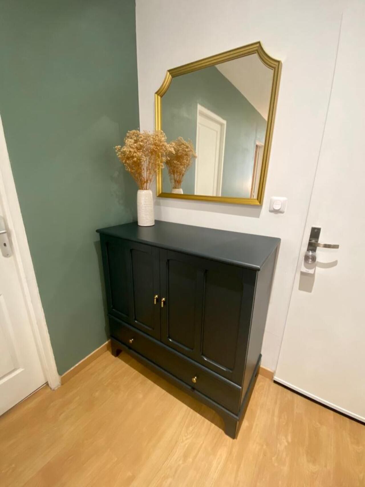 Een donkere houten kast met een witte vaas met gedroogde bloemen erop, een grote spiegel met gouden lijst, en een witte deur met zilveren knop.