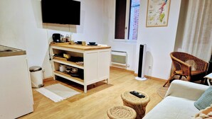 Un salon moderne comprenant un canapé, un fauteuil en osier, une petite table, un coin cuisine avec un meuble blanc, un téléviseur et une fenêtre donnant sur un immeuble.
