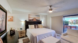 A bedroom with a bed, a ceiling fan, a nightstand, a chair, a window with a view, and artwork on the walls. - Terrasol Beach Resort, Oceanfront 2BR Condo #106 (Cabo San Lucas)