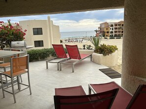 An outdoor patio with red chairs, a grill, and a view of the beach. - Terrasol Beach Resort, Oceanfront 2BR Condo #106 (Cabo San Lucas)