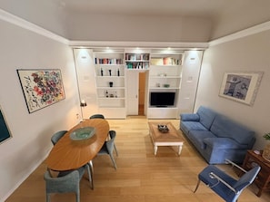 Ein gemütlicher Wohnraum mit Esstisch, Sofa, Fernseher und integrierten Regalen mit Büchern und Dekoration.