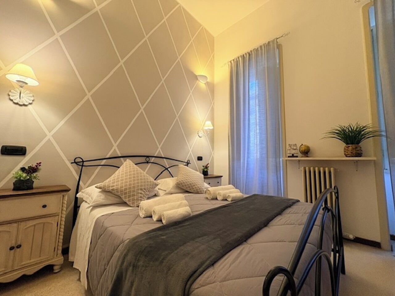 A neatly made bed with pillows and a blanket, a bedside table with a lamp, a radiator, and a shelf with a plant.