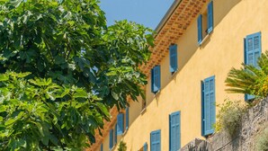 A cobblestone street leading to a yellow building with blue shutters and a stone wall. - Domaine de la Cascade Parfumée (Grasse)