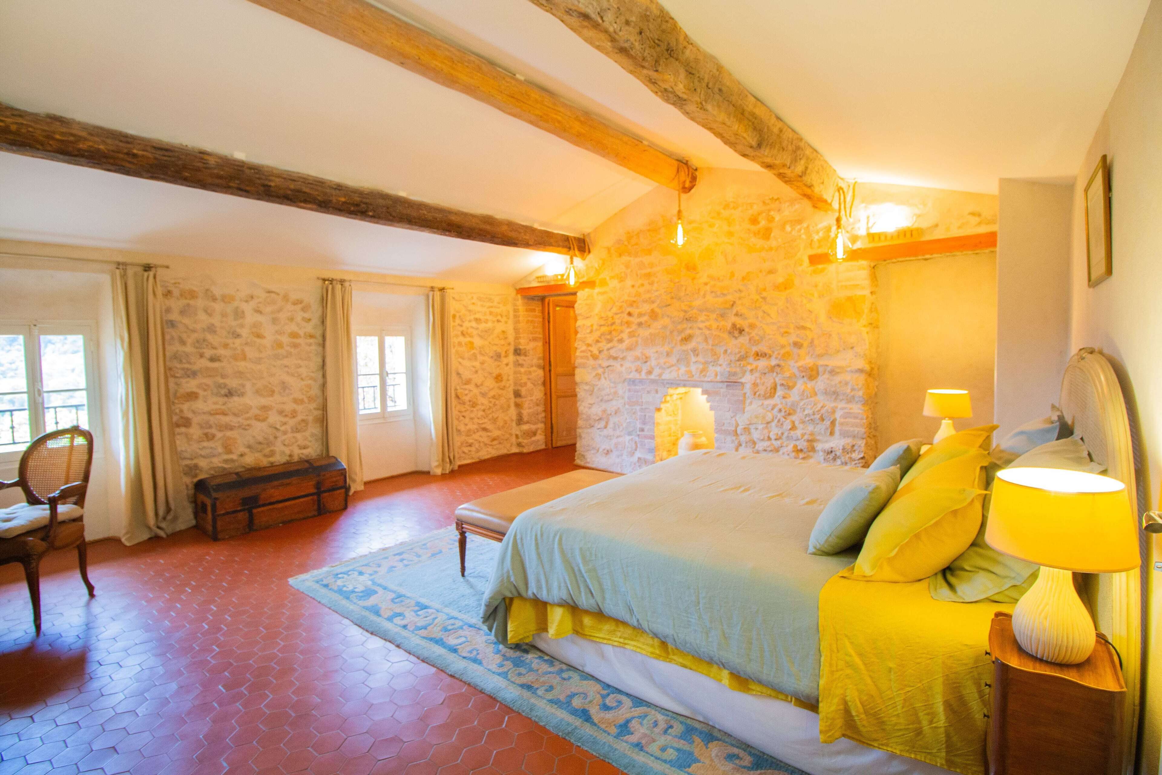 Un dormitorio con pared de piedra, viga de techo de madera, cama con ropa de cama amarilla, mesita de noche con lámpara y ventana con cortinas.