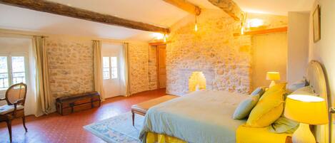 Un dormitorio con pared de piedra, viga de techo de madera, cama con ropa de cama amarilla, mesita de noche con lámpara y ventana con cortinas.