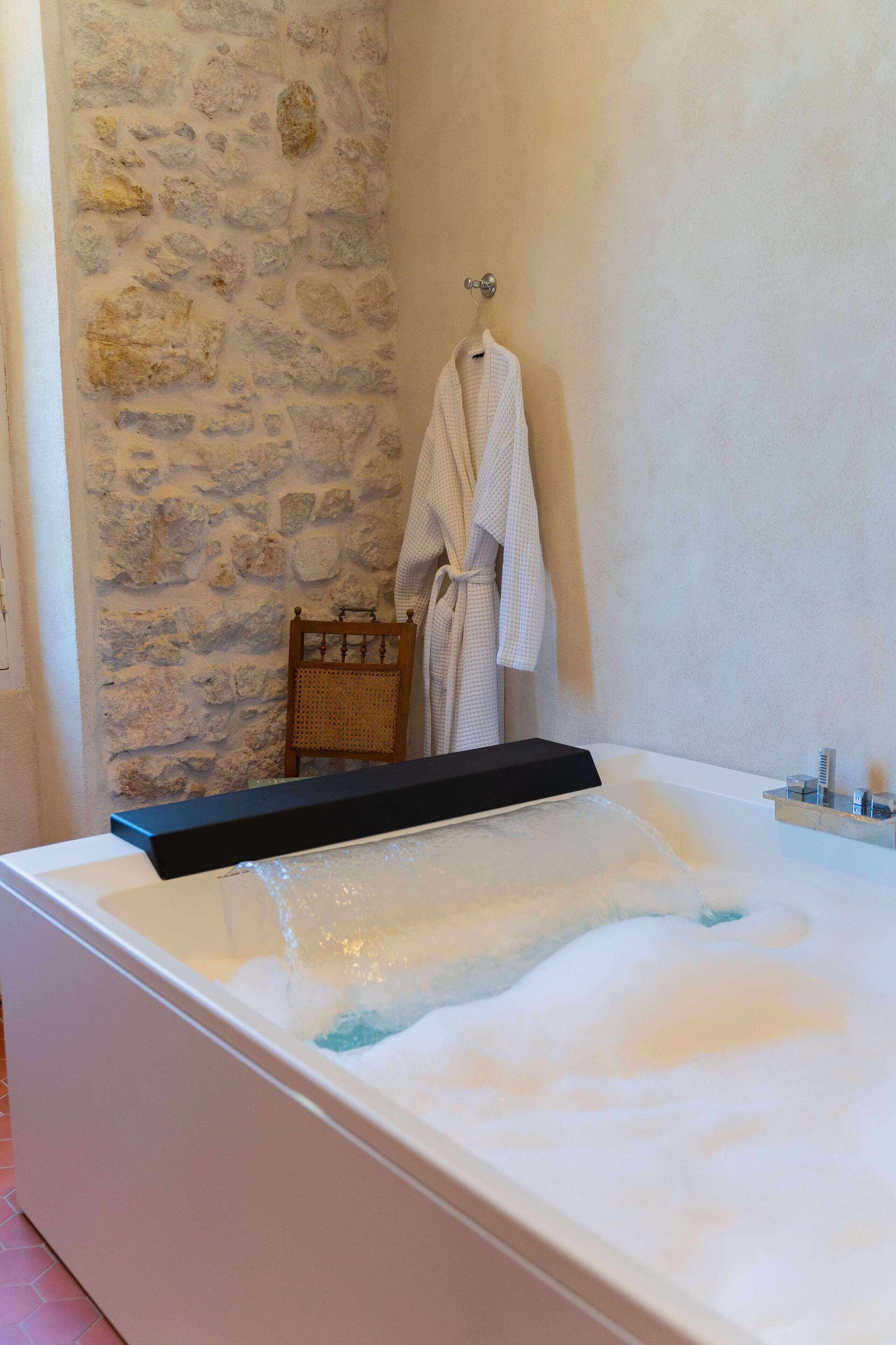 A modern bathroom with a stone wall, a bathtub with a black tray, and a wooden chair.