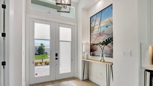 Et moderne interiør med en stor glasdør, der fører til en terrasse, et maleri af en strandscene, og et lille bord med en lampe.