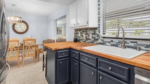 Une cuisine moderne avec un plan de travail en bois, des meubles de couleur sombre, un évier encastré et une fenêtre équipée de stores.