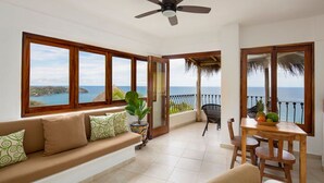 A balcony with a view of the sea, a sofa with patterned cushions, a dining table with chairs, and a ceiling fan.