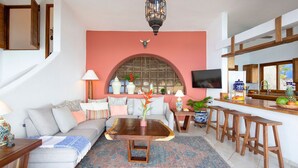 A living room with a sofa, coffee table, and bar stools. There is a TV mounted on the wall and a large window with a decorative arch.