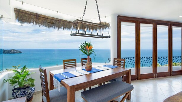 A wooden dining table with blue placemats, a vase, and chairs. A potted plant is on the left. The room has a view of the sea and a thatched ceiling.