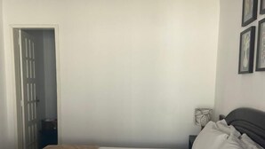 Ein ordentlich bezogenes Bett mit weißer Bettwäsche und Kissen, ein Nachttisch mit Lampe sowie eine Tür zu einem anderen Raum.