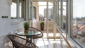 A balcony with a glass table and wicker chairs, potted plants, and a view of buildings.