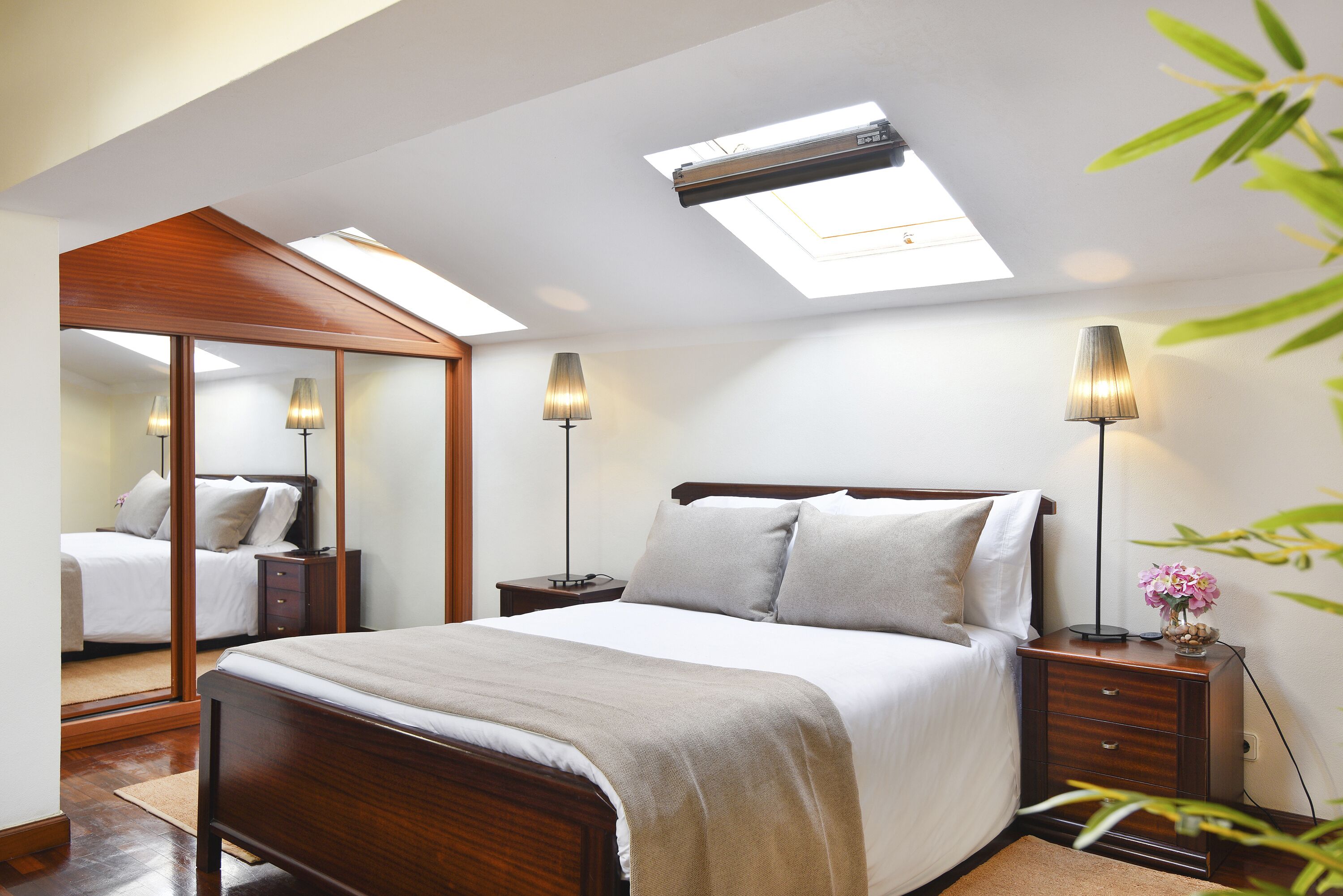 Un dormitorio con tragaluces, una cama con cabecera de madera, mesitas de noche con lámparas y un espejo.