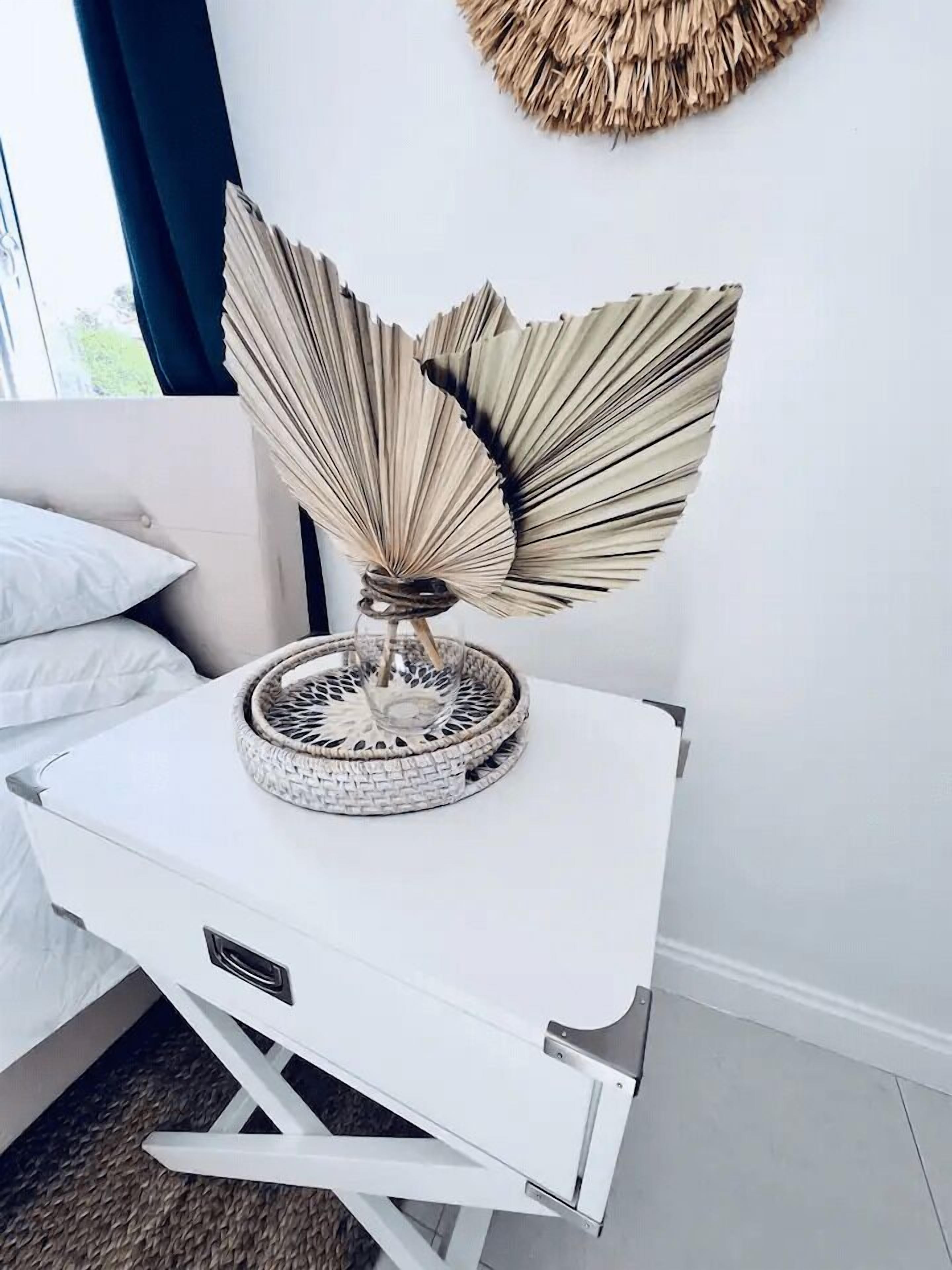 Une table pliante blanche, ornée d’un éventail décoratif en métal sur le dessus, est placée dans une pièce qui contient un lit et une décoration murale en fibres tressées.