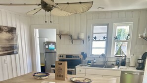 A kitchen with a central island, two blue bar stools, a ceiling fan, and a refrigerator. - The Beach Box is located in the heart of CB, walking distance to the boardwalk! (Carolina Beach)