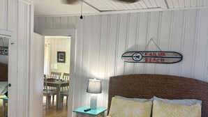 A bedroom with a bed, a ceiling fan, a nightstand, a lamp, and a sign on the wall. - The Beach Box is located in the heart of CB, walking distance to the boardwalk! (Carolina Beach)