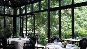 An indoor dining area with tables set for two, surrounded by large windows offering a view of lush greenery. - Arté Boutique Hotel (Tirana)