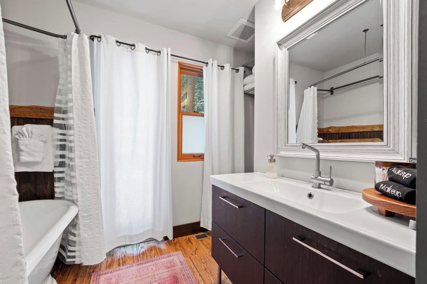 A bathroom with a shower curtain, a window, a sink with a mirror, and a vanity with a towel rack.
