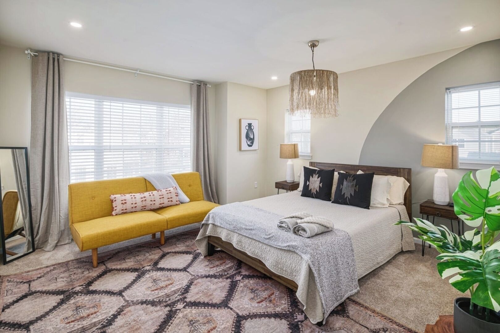 Een slaapkamer met een bed, een gele bank, een kamerkleed met dessin en een grote plant.