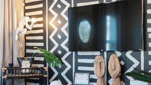 A modern living room with a large flat-screen TV mounted on a black and white patterned wall. There is a wooden coffee table with decorative items, a black console table with a plant, and a small wooden cabinet. - Imani Playce: Perfect location w/ Boho Vibes! (Atlantic City)