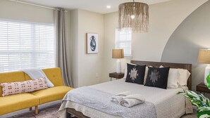 A bedroom with a bed, a sofa, a hanging light fixture, a bedside table, and a window with curtains. - Imani Playce: Perfect location w/ Boho Vibes! (Atlantic City)