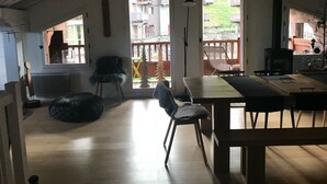 A spacious living area with a high ceiling, a dining table, chairs, a fireplace, and a view of a balcony with outdoor furniture.