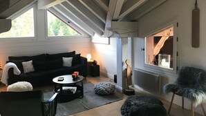 A cozy room with a dark-colored sofa, a small round table, and a chair. There is a window with a view of trees, and a wooden beam ceiling.