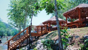 A wooden staircase leading to a thatched-roof structure by a river, surrounded by greenery.