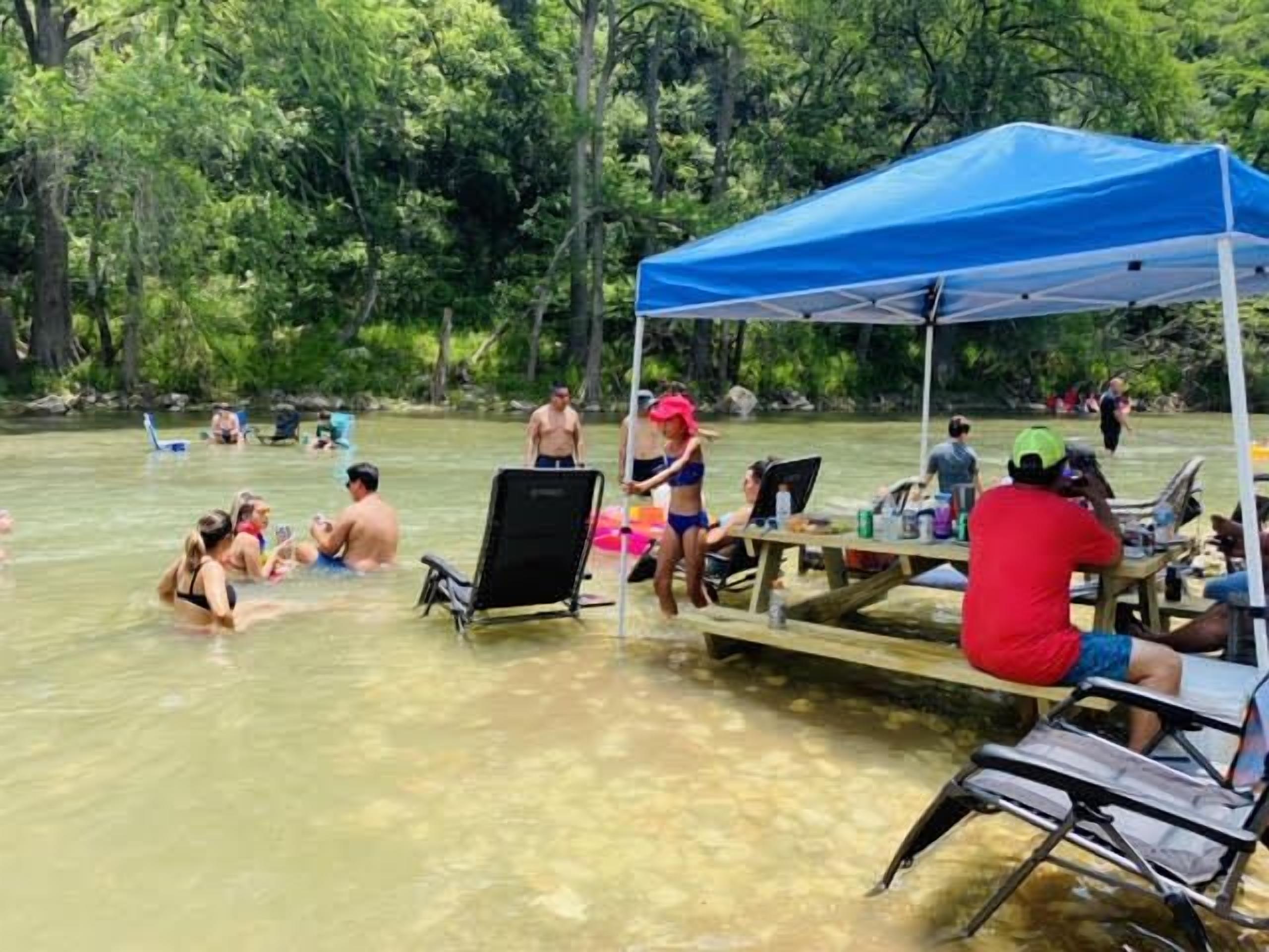 Un grupo de personas en un entorno natural bajo un dosel azul, algunas sentadas en mesas y otras en el agua.