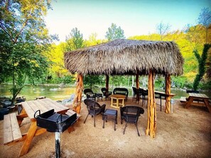 Un espacio para comer al aire libre con techo de paja, mesas de picnic y sillas, rodeado de árboles y un río.