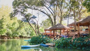 A serene riverside setting with thatched-roof huts, wooden benches, and small boats.