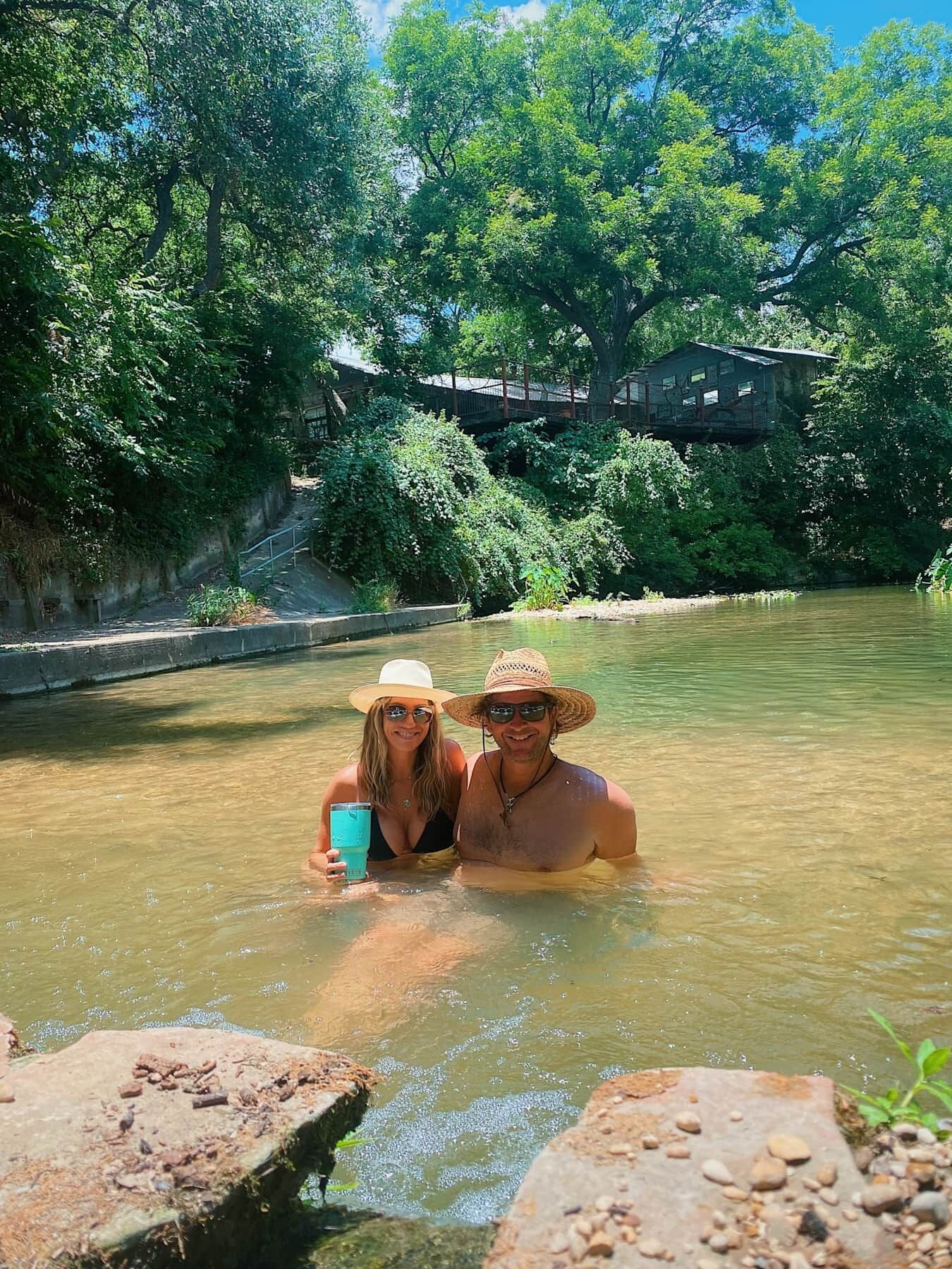 Two people in a river with a bridge and greenery in the background.