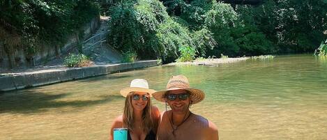Two people in a river with a bridge and greenery in the background.