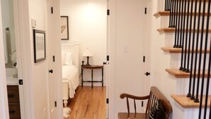 Un couloir au plancher en bois, un escalier avec des balustrades noires et une chaise dotée d’un coussin à motifs.