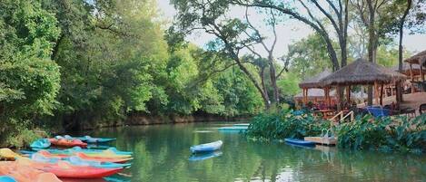 A calm river with colorful boats docked on the left, surrounded by lush greenery and a thatched-roof structure on the right.